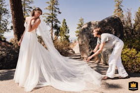In South Lake Tahoe, California, a wedding photographer captures a serene moment. The image shows an attendant adjusting the train of the bride's dress, bathed in warm sunshine during the outdoor wedding.